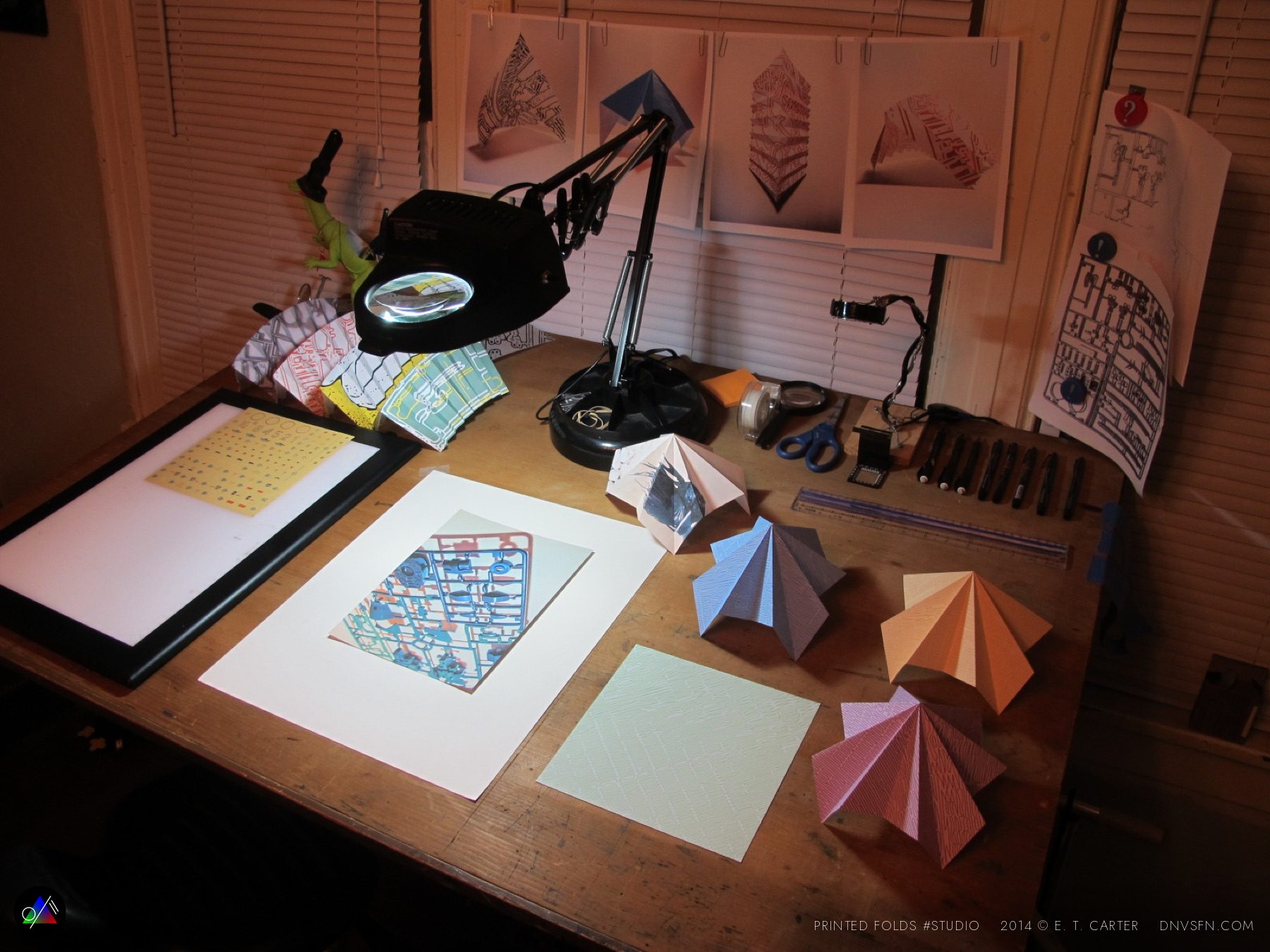 The work desk at night, prints pinned to the blinds, magnifier lamp and cutters out, folded forms drying.